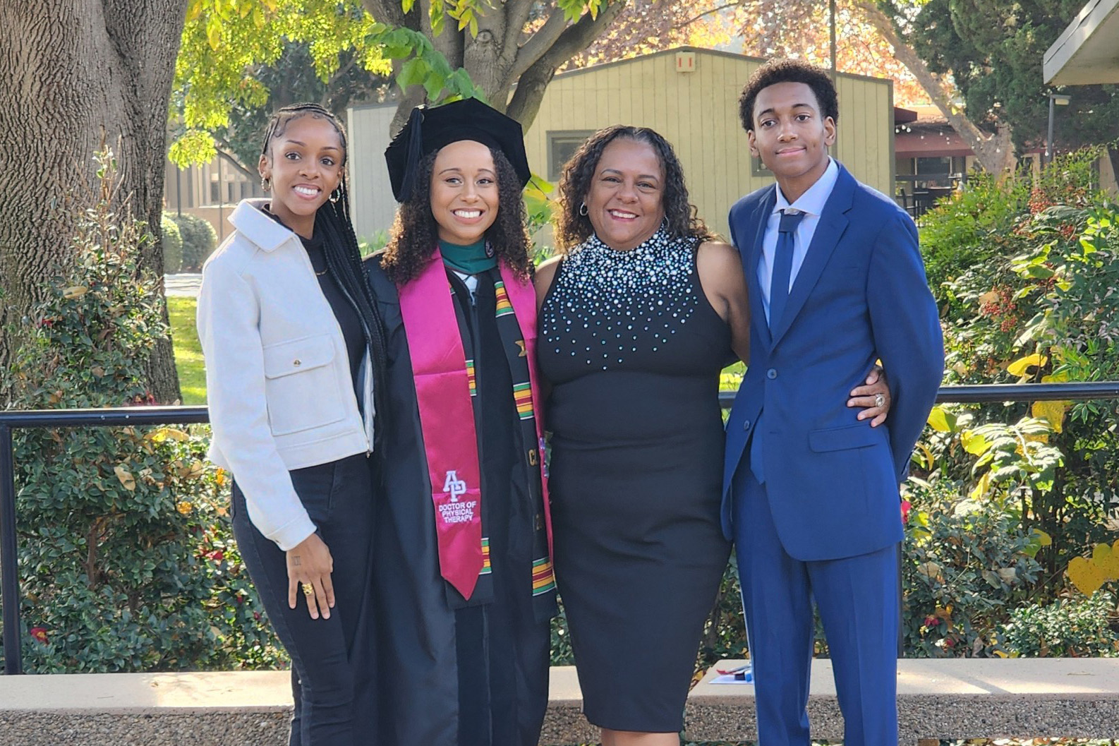 Genee Johnson A smiling family of four celebrating a graduation outdoors. In the center, a young woman wearing a black graduation gown and a pink "Doctor of Physical Therapy" sash stands next to an older woman in a black dress with a beaded neckline. They are flanked by another young woman in a white jacket and a young man in a blue suit.