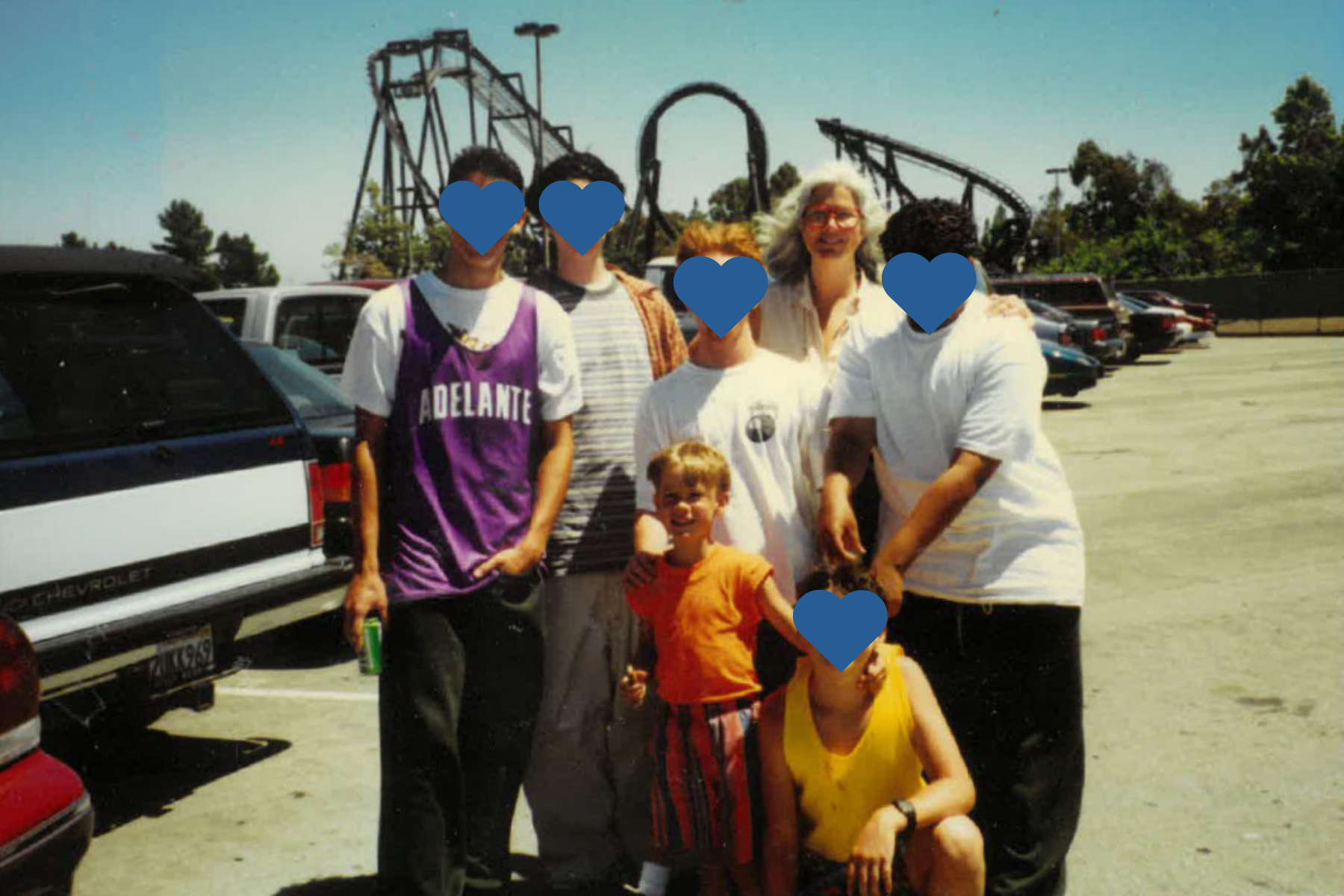 A group of six people posing for a photo in a parking lot, with a large rollercoaster in the background on a sunny day. A woman with glasses is smiling with five young people, including one younger child. One of the youth wears a purple jersey that reads "ADELANTE." The image highlights a family moment during a day of recreation. The group and woman show a joyful connection. The background includes a large parking lot, multiple cars, and a massive steel rollercoaster with multiple tracks.