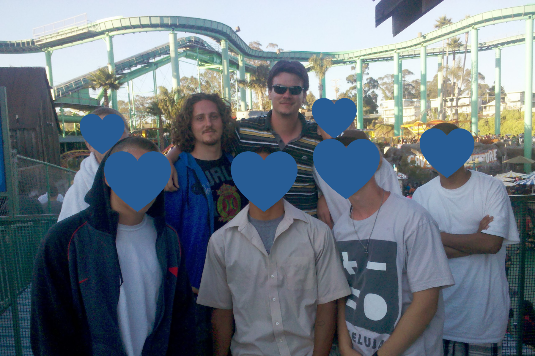 A group of seven young men pose together outdoors at an amusement park on a bright, sunny day. A tall green roller coaster track arches high above them in the background against a clear sky. Two young men in the back row are unmasked, one wearing sunglasses and a striped polo shirt, and the other with curly hair in a blue zip-up hoodie. Five other group members stand closely together around them in casual attire.