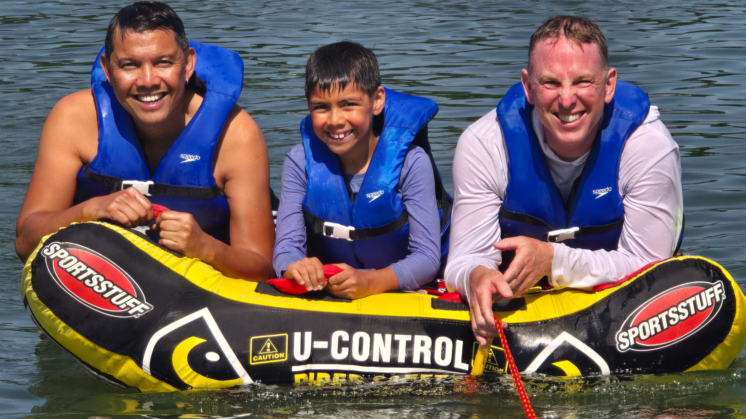 34652 A family of two fathers and their young boy wearing blue life jackets, smiling while riding together on a yellow inflatable water tube on a lake.