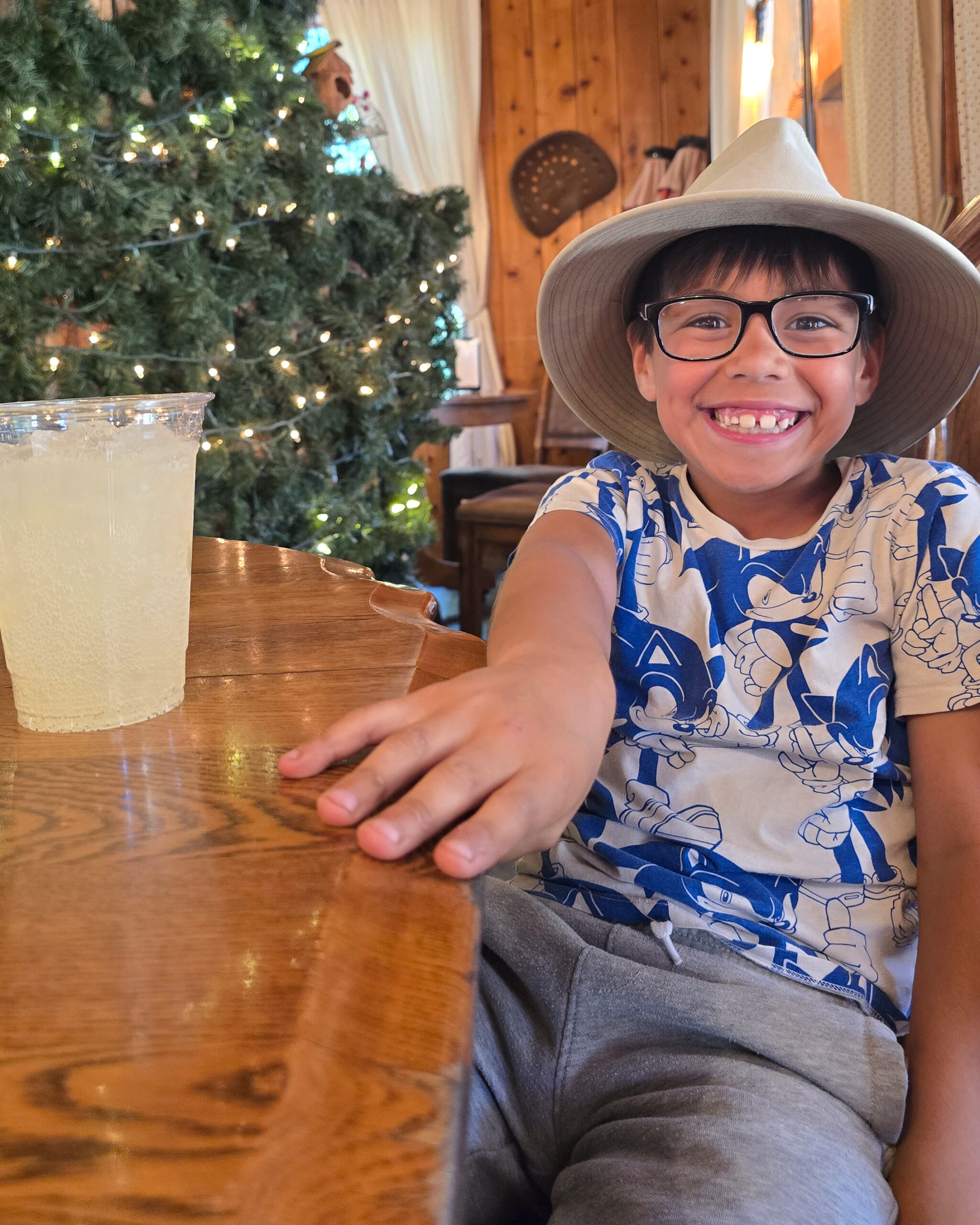34656 A smiling young boy wearing a wide-brimmed hat, glasses, and a blue Sonic the Hedgehog shirt sitting at a wooden table next to a cold drink, with a lit Christmas tree in the background.