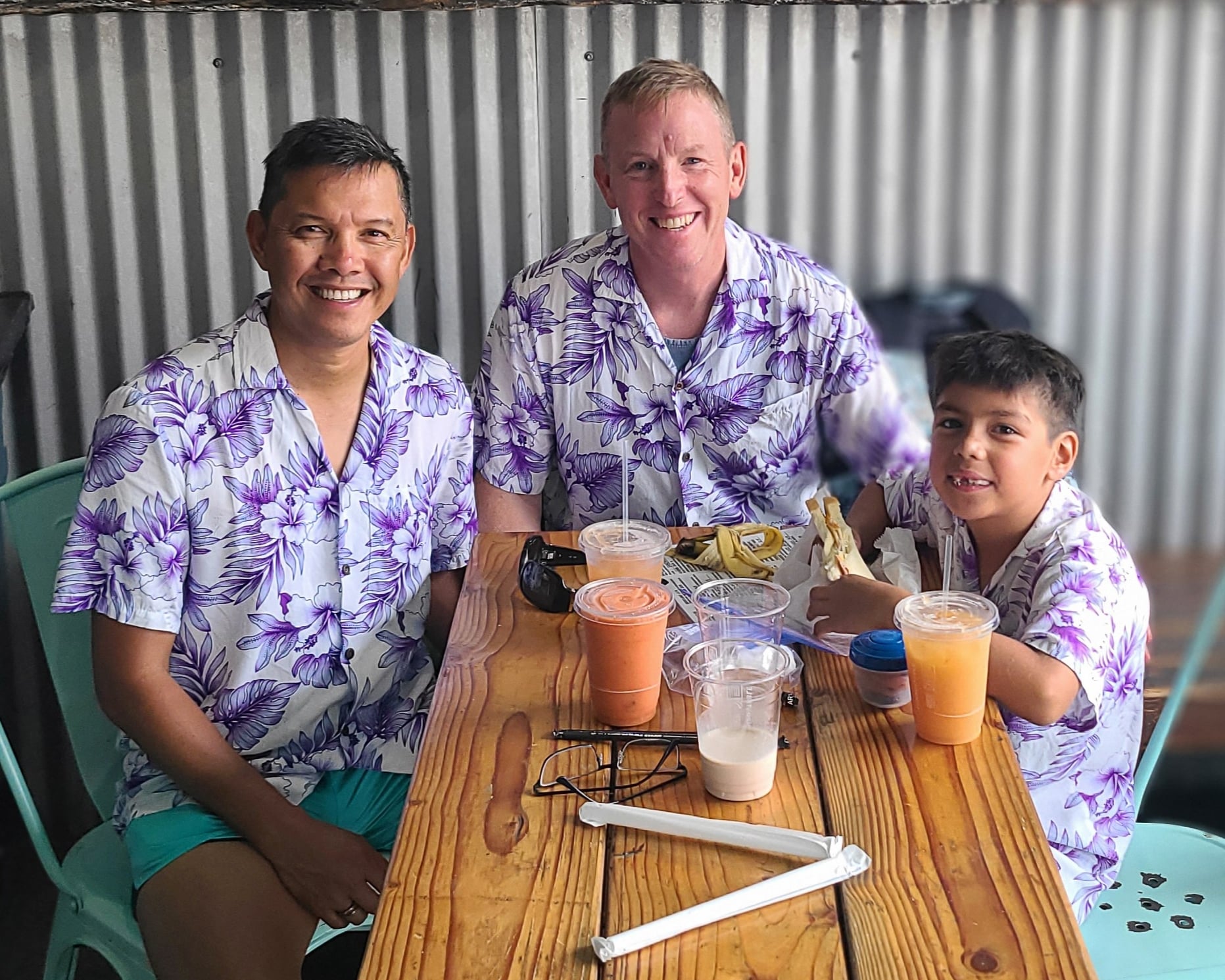 34657-2 wo smiling dads and their young son sitting together at a wooden table enjoying drinks and food while wearing matching purple floral tropical shirts.