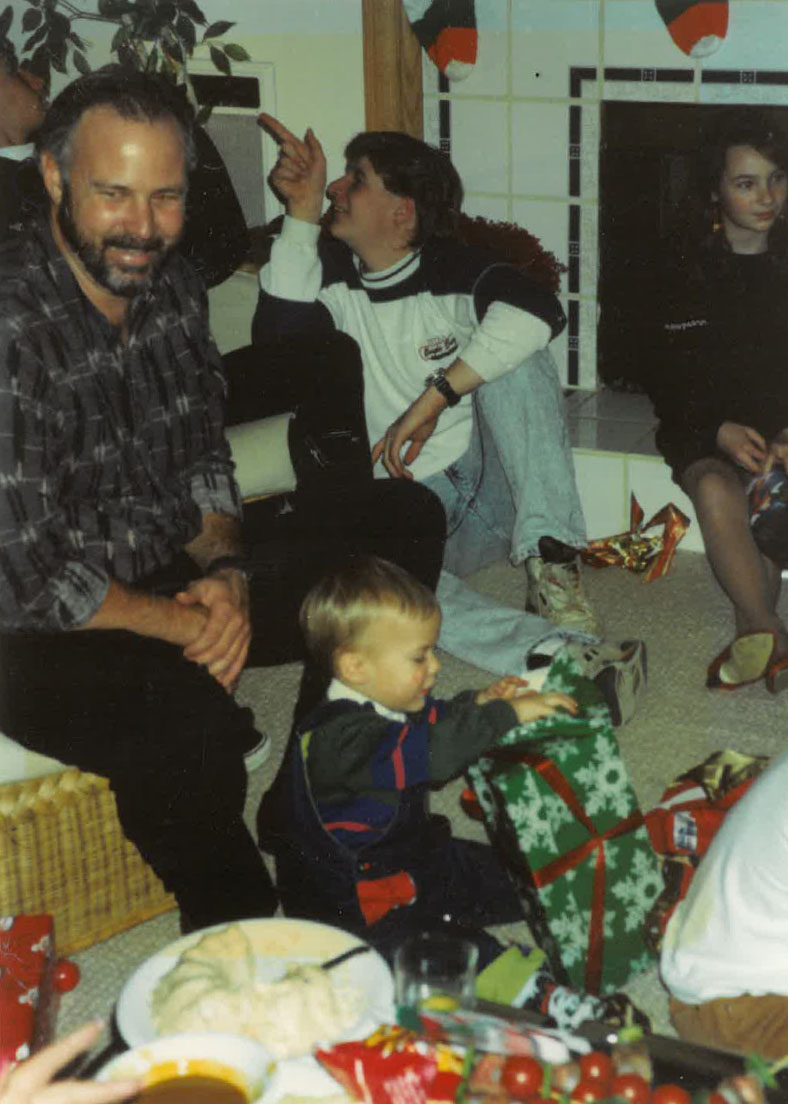 A vintage photo of a family gathered in a living room around a fireplace during a Christmas gift exchange. In the foreground, a young toddler sits on the carpet opening a green gift wrapped with white snowflake paper. A bearded man sits beside him smiling, while two older children sit on the floor near the hearth, with holiday stockings hanging above. A spread of food and snacks is visible in the bottom foreground.